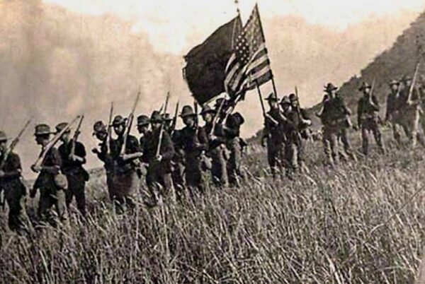 Column of U.S. infantry with Stars and Stripes advancing through tall grass in Lanao, 1902.