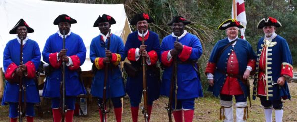 Men of African descent in blue 18th-century uniforms holding muskets during a Fort Mose reenactment.